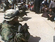 U.S. soldiers kneel and lower their weapons, a gesture to show they mean no threat; some in the crowd follow suit and sit down.